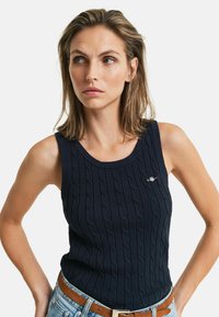 Navyblauwe gebreide tanktop met een kabelpatroon, ronde halslijn en een klein logo op de borst. Gedoord met lichtblauwe denim shorts en een bruine riem.