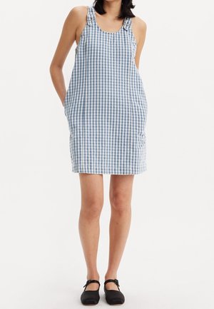 Femme portant une robe sans manches à carreaux bleus et blancs avec des bretelles à boucle et des ballerines Mary Jane noires, debout les mains derrière le dos.