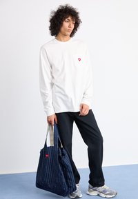 White long-sleeve shirt with a red heart emblem on the pocket, paired with black pants and a navy patterned tote bag. Gray sneakers.