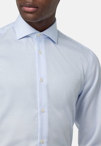 Camisa de vestir rayada en azul claro con cuello abotonado, fila sencilla de botones y una textura suave. Las rayas son paralelas y delgadas.