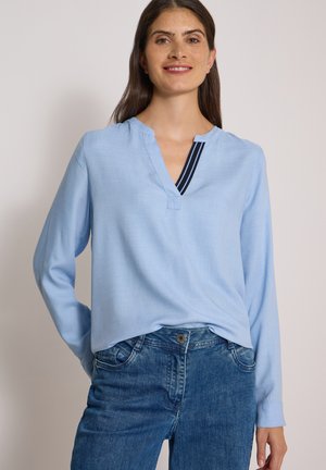 Femme aux longs cheveux bruns souriante, portant un chemisier bleu clair à manches longues avec un détail col en V rayé et un jean bleu en denim.