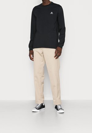 Mand iført sort langærmet Converse-trøje, beige bukser og sorte høje sneakers, stående mod en ensfarvet hvid baggrund.