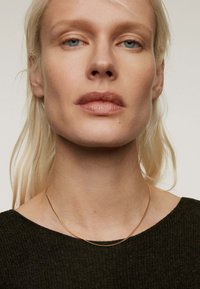 Collar de cadena de oro llevado sobre un suéter oscuro y texturizado. El collar tiene un diseño delicado y liso con una ligera curvatura.