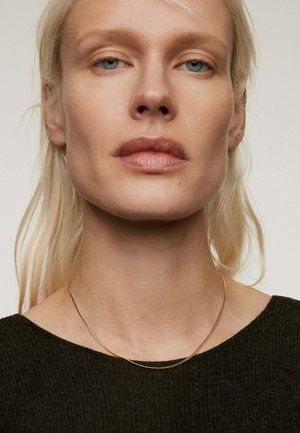 Collar de cadena de oro llevado sobre un suéter oscuro y texturizado. El collar tiene un diseño delicado y liso con una ligera curvatura.