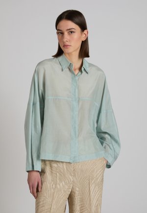 Młoda kobieta o prostych brązowych włosach ubrana w luźną, jasnoniebieską koszulę na guziki i beżowe spodnie o fakturze, stoi na tle jednolitego szarego tła.