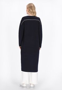 Marineblaues langärmliges Kleid mit weißen Akzentnähten, lockerer Passform, seitlichen Schlitzen und hohem Seitensaum. Kombiniert mit weißen weiten Hosen und Turnschuhen.