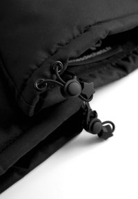 Musta tyynyverhoiltu vaate, jossa on sileä kangas, kiristysnyörit, joissa on pyöreät ja tähti muotoiset muoviset soljet säätämistä varten.