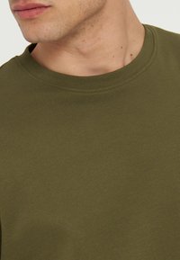 Gros plan d'un homme portant un t-shirt vert olive à col rond, montrant l'encolure et le bas du visage avec un teint clair.