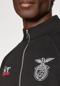 Black zip-up sweatshirt with high collar, featuring white embroidered eagle logo and text. Material appears smooth and slightly textured.