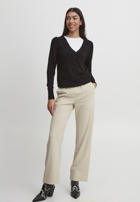 Schwarzes Langarmshirt mit V-Ausschnitt und Knopfdetails, kombiniert mit cremefarbenen weit geschnittenen Hosen und schwarzen Stiefeletten mit weißen Akzenten.