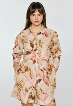 Kobieta w beżowej koszuli na guziki i dopasowanych szortach z różowym i brązowym wzorem liści, stojąca z rękami w kieszeniach.