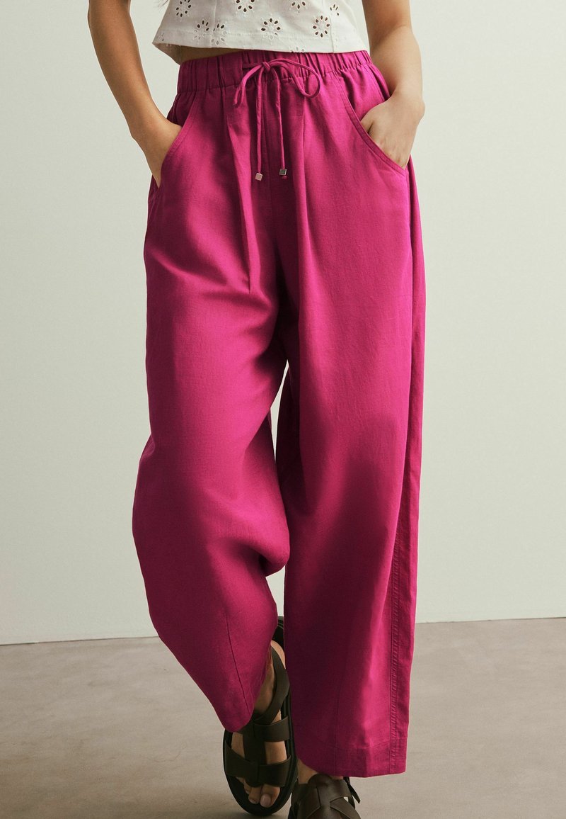 Donna che indossa pantaloni larghi magenta con vita con coulisse e tasche, abbinati a sandali neri, in piedi su un pavimento grigio.