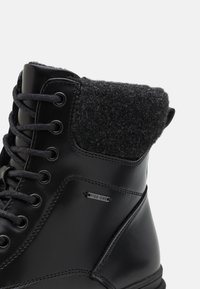 Botas negras hasta el tobillo con un acabado de cuero suave y un cuello de fieltro gris, que presentan cordones negros, ojales de metal y un pequeño logo plateado.