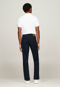 Pantaloni navy con vestibilità dritta, cuciture dettagliate, tasche posteriori e una toppa in pelle marrone in vita. Abbinati a una maglietta polo bianca.