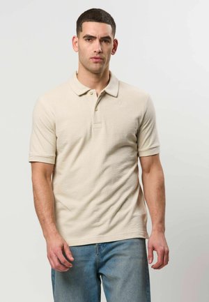 Homme portant un polo beige à manches courtes et un jean bleu, debout devant un fond gris clair uni.