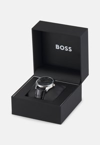 Reloj con correa de cuero negro en una caja negra, con un caso plateado y una esfera negra con un patrón texturizado y acentos dorados. Logo de BOSS presentado en el interior.