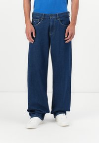 Muškarac u širokim tamnoplavim trapericama i bijelim tenisicama stoji nasuprot jednostavne bijele pozadine.