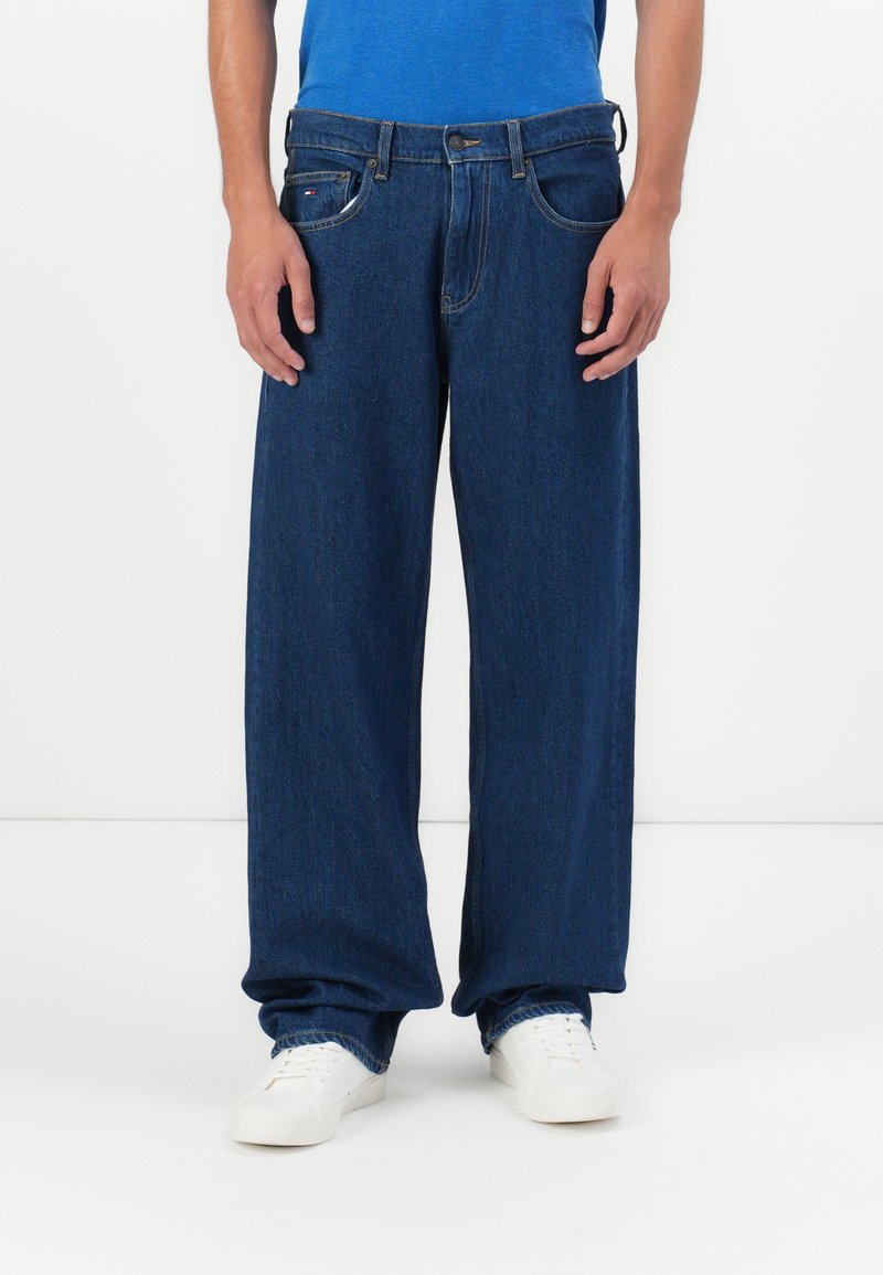 Muškarac u širokim tamnoplavim trapericama i bijelim tenisicama stoji nasuprot jednostavne bijele pozadine.