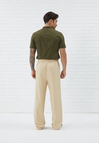 Olivengrøn kortærmet skjorte med rygstykke, parret med beige brede bukser med elastisk talje, præsenteret mod en hvid murstensbaggrund.