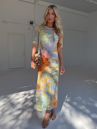 Vestido comprido justo em tecido multicolorido com padrões abstratos de folhas. Mangas curtas, decote redondo, combinado com sandálias com tiras e uma carteira de palha.