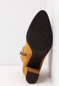 Bottine en daim marron avec semelle en caoutchouc noire et talon bloc, vue de dessous avec fermeture éclair latérale visible sur fond blanc.