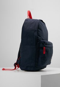 Námořnický batoh s červenými akcenty, textilní materiál, zakřivený tvar, boční kapsa se zipem a nášivka s logem na přední straně.