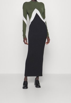 Tall person wearing a long, fitted dress with green, white, and black geometric color blocks paired with black heeled loafers.