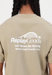 T-shirt khaki con logo stampato bianco "ReplayGoods" e testo "Les Tenues des Sportifs Monte-Carlo Country Club" sul retro.