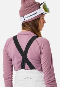 Paarse geribbelde lange mouw top met zwarte verstelbare bandjes met de tekst "SWDEMO", gecombineerd met witte bretelpantalons en een gebreide roze beanie.