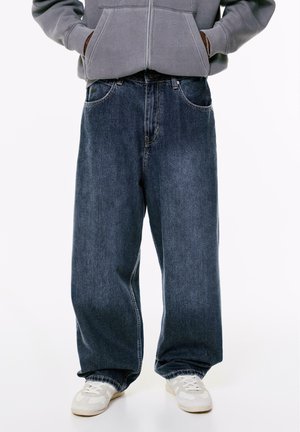 Jeans in denim blu scuro a gamba larga con un classico design a cinque tasche e cuciture sottili. Abbinati a sneaker di colore chiaro.