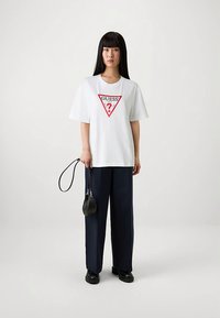 Wit katoenen T-shirt met een rood driehoekig "GUESS?"-logo in het midden, gecombineerd met marineblauwe broek met wijde pijpen en zwarte schoenen en handtas.