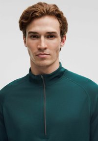 Jaunas vyras su šviesiai rudais plaukais, vilkintis tamsiai žalią užtrauktuką, žiūri tiesiai į priekį prieš paprastą šviesią foną.