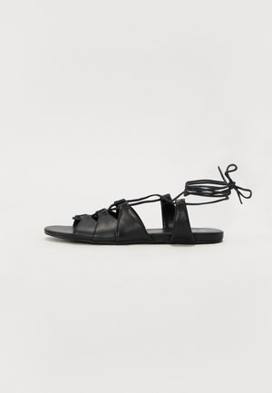 Sandalias de cuero negro con suela plana, correas entrecruzadas y lazos en el tobillo, con un diseño minimalista de punta abierta.