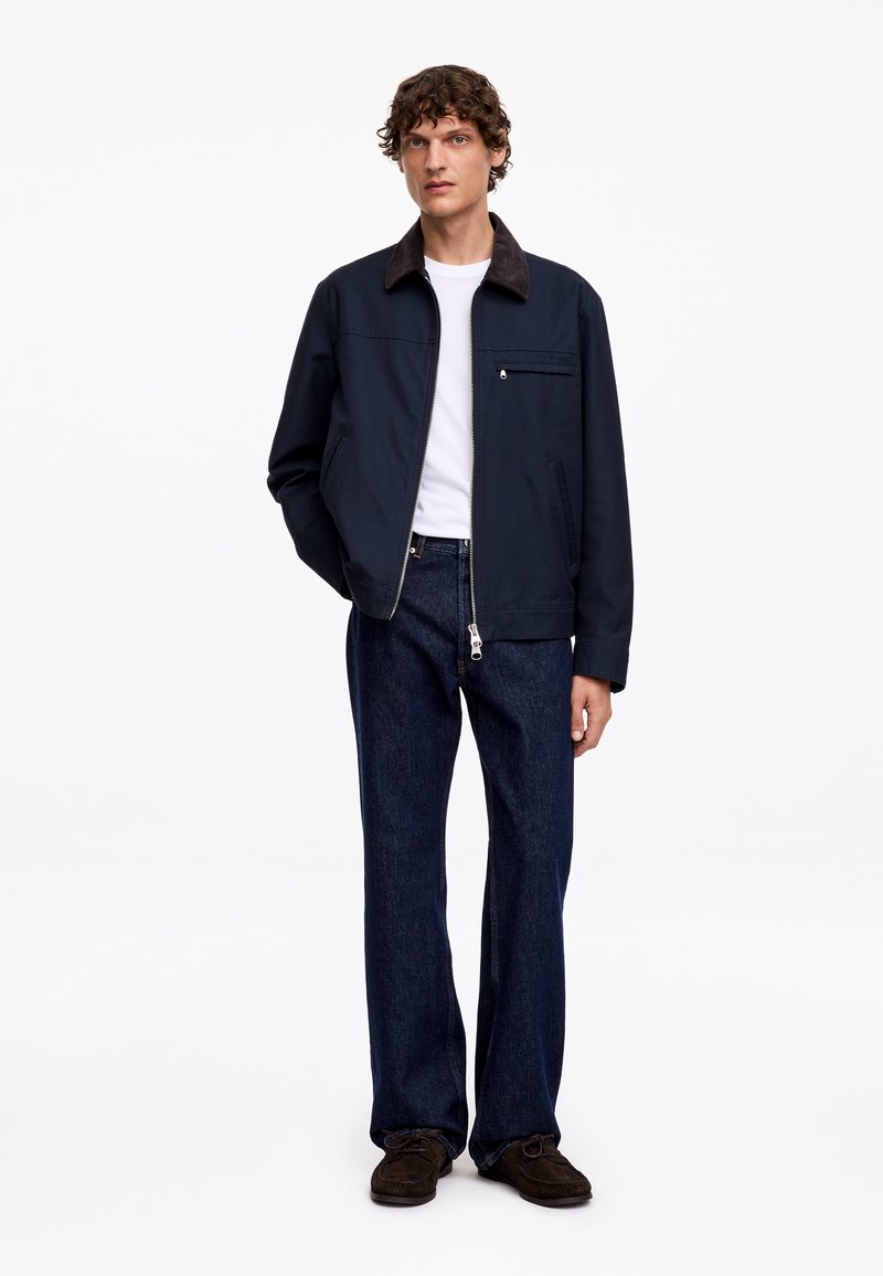 Homme portant une veste bleu marine, un t-shirt blanc, un jean bleu foncé et des chaussures marron, debout avec une main dans la poche, sur un fond blanc.