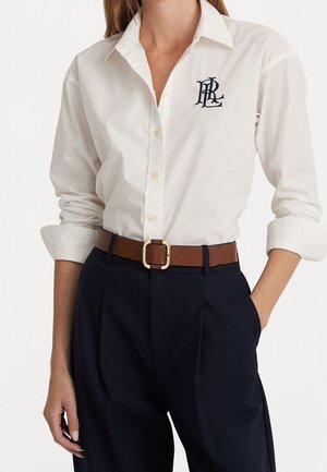 Femme portant une chemise blanche à boutons avec les manches retroussées, un pantalon bleu marine et une ceinture marron avec une boucle dorée, debout avec une main dans la poche.