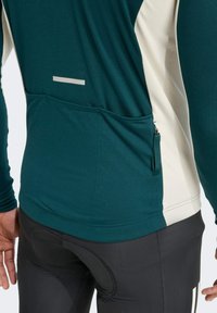 Langärmliges Sportoberteil in Türkis und Creme mit einer Reißverschlusstasche, glatter Textur und reflektierendem Streifen auf dem Rücken. Schwarze, enge Hose.