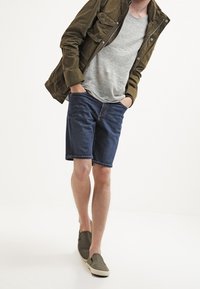 Vaatteet rento asu, jossa on harmaa t-paita, oliivinvihreä takki, siniset farkkushortsit ja harmaat liukuhihnat, esiteltynä valkoista taustaa vasten.