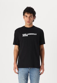Juodas medvilninis marškinėliai su apvaliu kaklu, trumpomis rankovėmis ir balta „KARL LAGERFELD JEANS“ logotipu, spausdintu per krūtinę.
