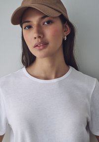T-shirt bianca in cotone con maniche corte, scollatura rotonda, indossata sotto un cappello marrone. Design semplice, texture liscia, senza pattern o dettagli visibili.