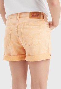 Pantalones cortos de mezclilla en naranja claro, con dobladillos enrollados, bolsillos traseros con detalles de costura y una etiqueta de marca marrón en la cintura.