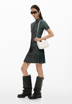 Frau in einem kurzärmeligen, schwarz-grün gestreiften Kleid, schwarzen kniehohen Stiefeln, Sonnenbrille und mit einer weißen Schultertasche.