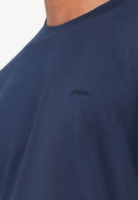 Κοντινό πλάνο ενός ατόμου που φοράει ένα ναυτικό μπλε T-shirt με στρογγυλή λαιμόκοψη και ένα μικρό, σκούρο κεντητό λογότυπο στο στήθος.