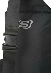 Bolsa negra con tejido texturizado, que presenta una correa robusta y un broche de plástico, con un logotipo plateado impreso en la parte frontal.