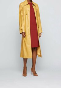 Trench-coat en cuir jaune avec col et boutons sur le devant, porté par-dessus une robe en maille rouge, associé à des chaussures à talons beiges.