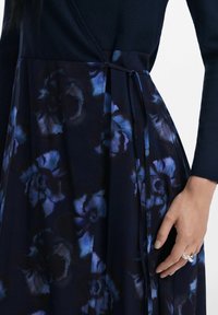 Vestido envolvente azul marino con una parte superior texturizada y una falda de estampado floral en azul oscuro y púrpura, con detalles de lazo y plisados.