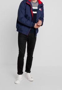 Marineblaue Zip-Jacke mit roten Akzenten, mit Kapuze, elastischen Bündchen und einem Logo-Patch. Über einem gestreiften Pullover und schwarzen Jeans getragen.