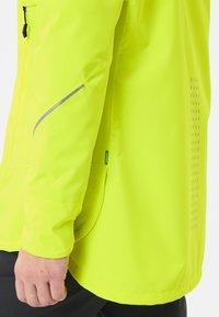 De linkerarm van een man draagt een felgele jas met een reflecterende strip op de mouw en een klein groen "groenvorm" label.