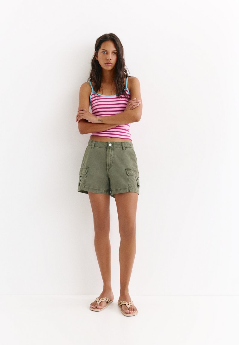 Femme debout, les bras croisés, portant un débardeur rayé rose, un short cargo olive et des sandales beiges sur fond blanc.
