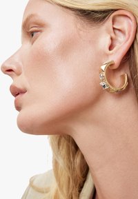 Boucles d'oreilles en or en forme de cercle avec une surface texturée, ornées de trois pierres rectangulaires transparentes. Le design complexe et la finition polie mettent en avant l'élégance.
