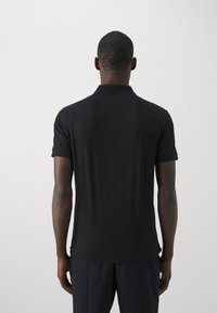 Man die met zijn rug naar de camera staat in een eenvoudig zwart T-shirt met korte mouwen en donkere broek, tegen een effen lichte achtergrond.